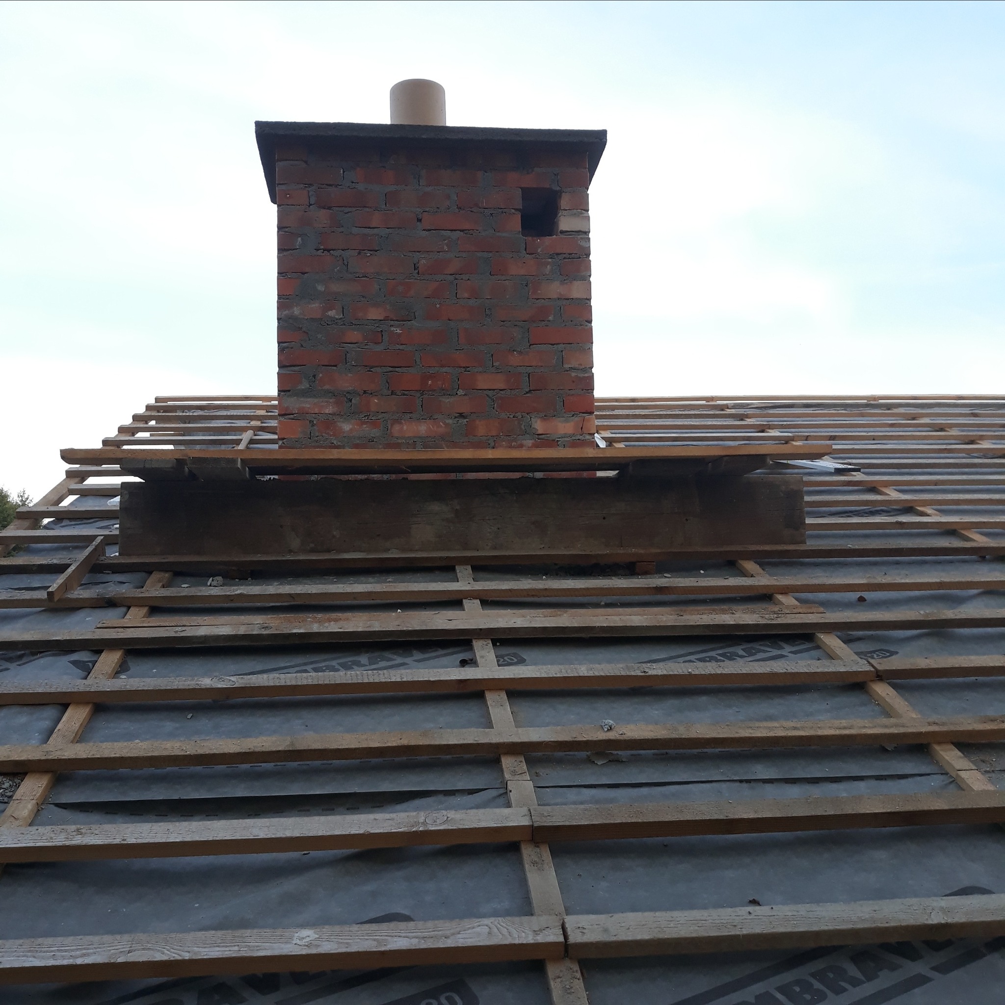 Ceglany komin na dachu z drewnianą konstrukcją i membraną dachową w trakcie budowy.