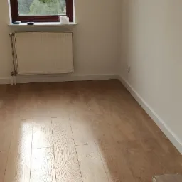 Jasne panele podłogowe ułożone w pustym pokoju z oknem i grzejnikiem, widoczny fragment opakowania paneli w rogu.