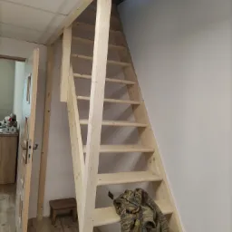 Surowe drewniane schody strychowe z widocznym oknem dachowym, częściowo otynkowaną ścianą i starą, drewnianą stolarką drzwiową.