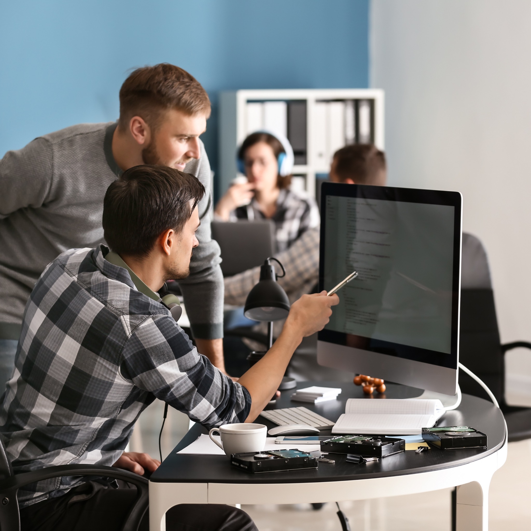 Zespół programistów analizuje kod na monitorze, na biurku leżą dyski twarde, notatnik i filiżanka kawy.