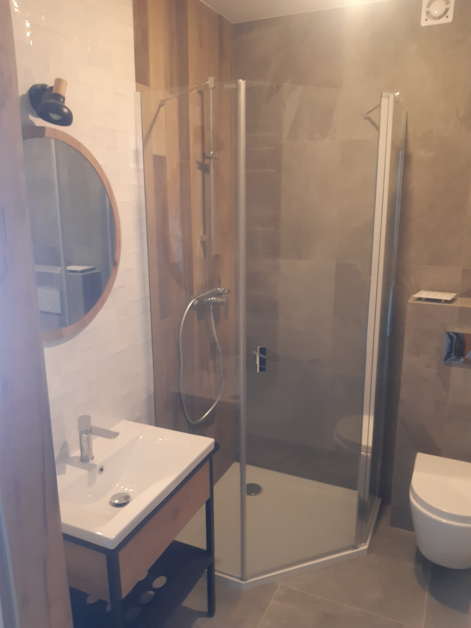 Nowoczesna łazienka z kabiną prysznicową, umywalką na szafce z półką i okrągłym lustrem z nietypowym oświetleniem na ścianie z białymi płytkami.