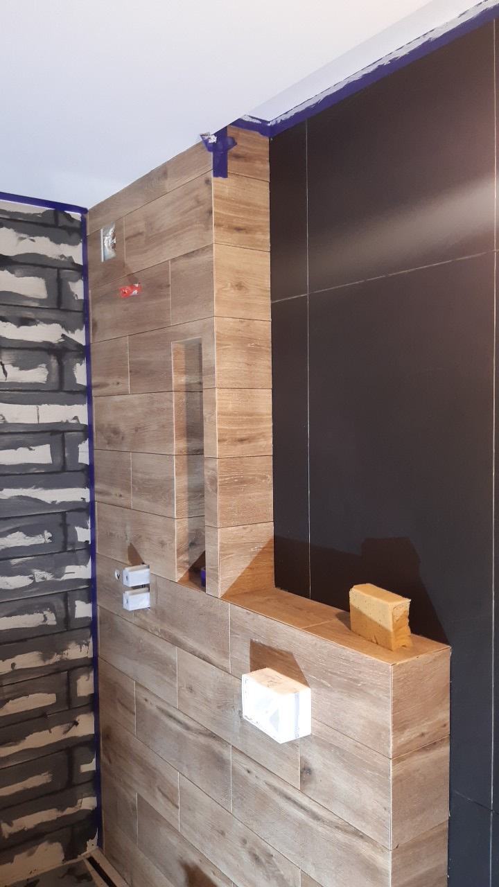 Ściana łazienki w trakcie wykańczania: brązowe płytki imitujące drewno na ścianie z wnęką, obok ściana z ciemnych, dużych płytek, widoczne niebieskie taśmy malarskie oraz fragment ściany z szarymi...