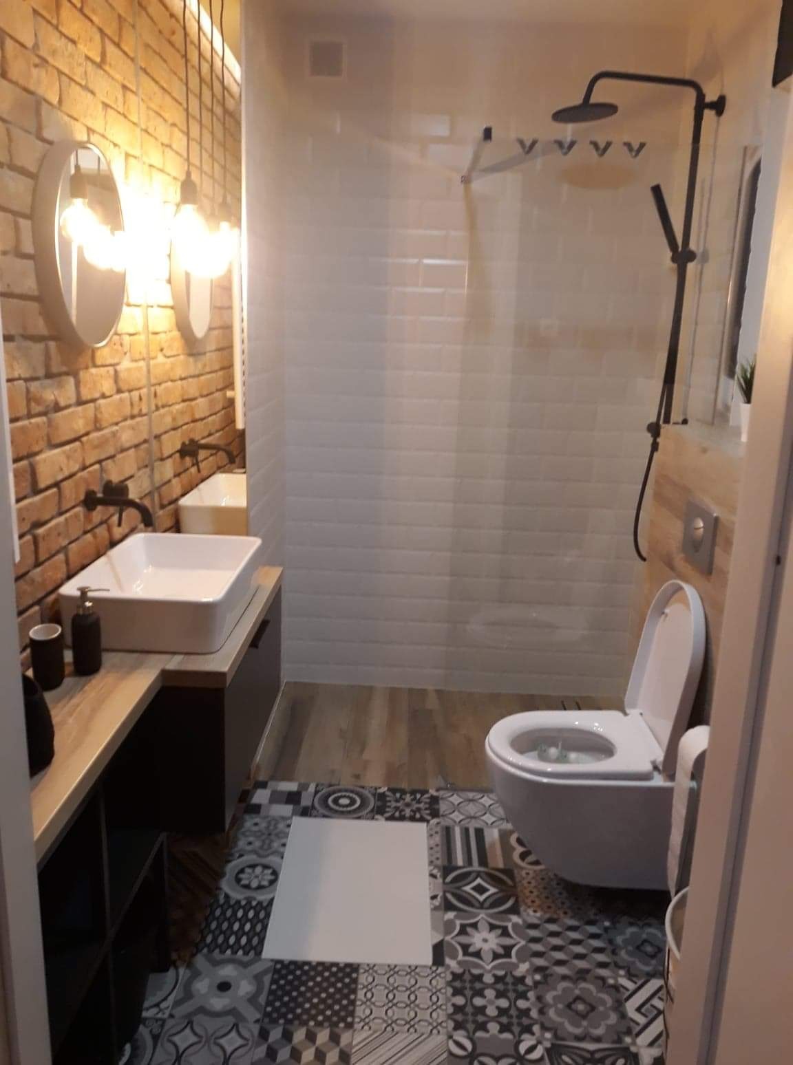Nowoczesna łazienka z ceglaną ścianą, wiszącą umywalką, kabiną prysznicową typu walk-in i wzorzystą podłogą z płytek.