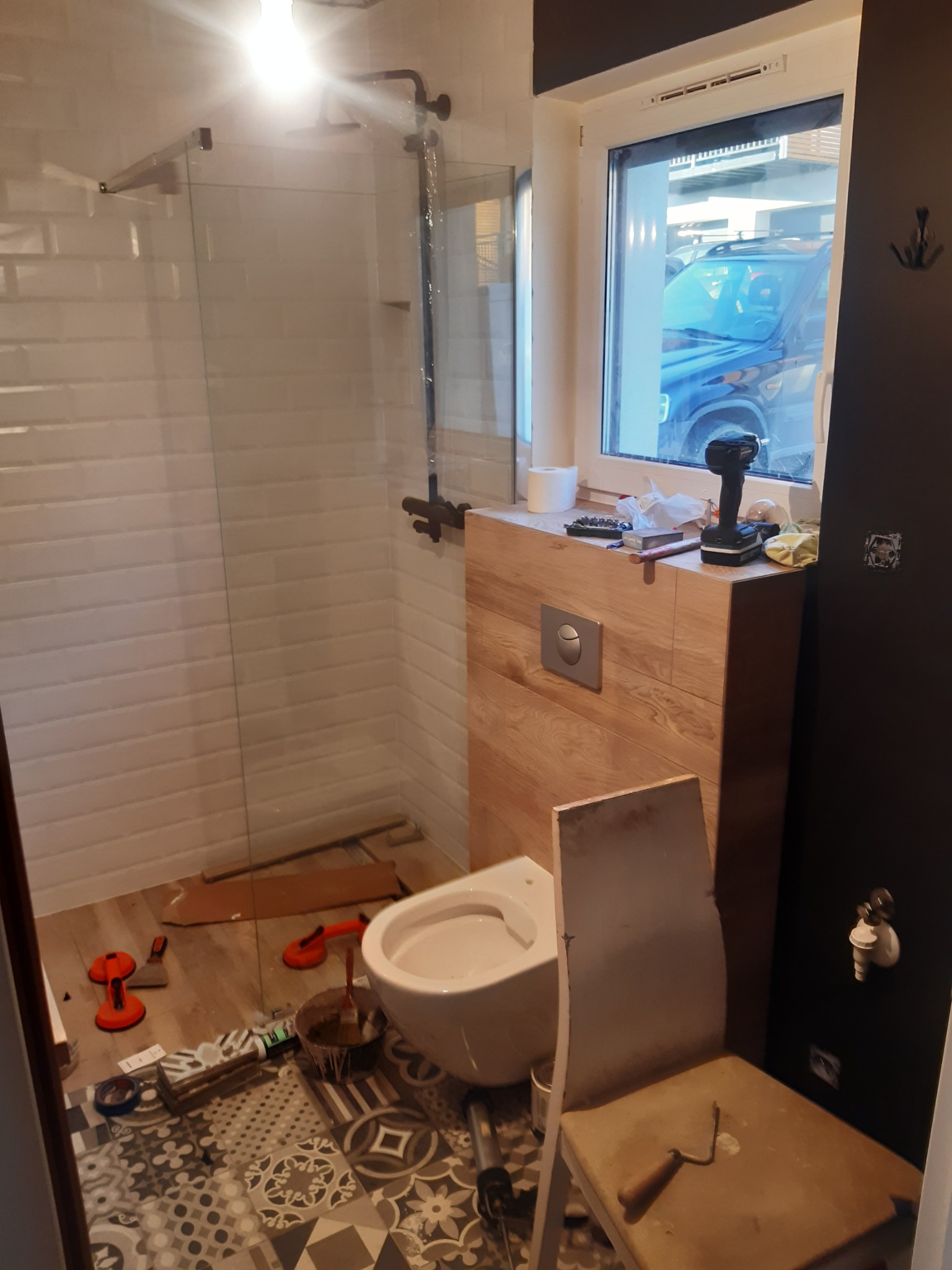 Remontowana łazienka z widoczną kabiną prysznicową z białymi płytkami, toaletą, drewnianą zabudową, oknem z widokiem na samochód oraz narzędziami i akcesoriami remontowymi. Na podłodze położone są...