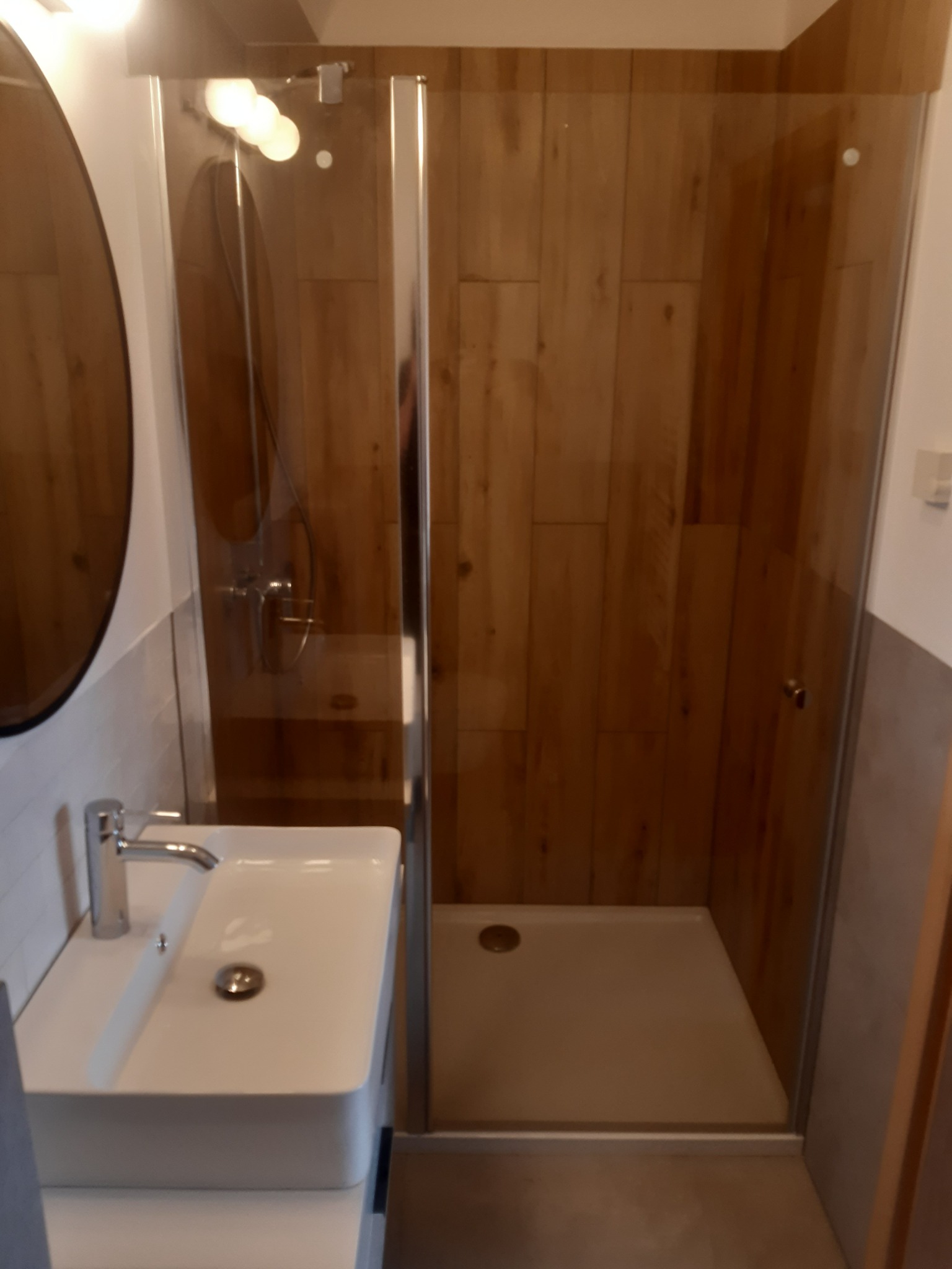 Nowoczesna łazienka z kabiną prysznicową typu walk-in z drewnianymi panelami, białą umywalką nablatową i okrągłym lustrem.