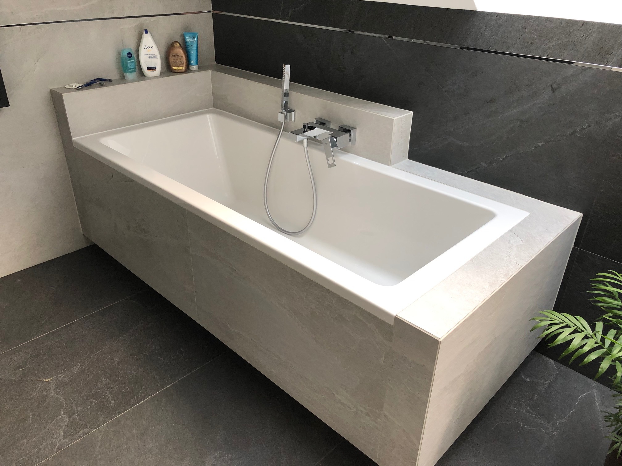 Nowoczesna wanna obudowana jasnoszarą płytką imitującą kamień, z chromowaną armaturą i półką na kosmetyki, na tle ciemnoszarej ściany z płytek i zielonej rośliny.