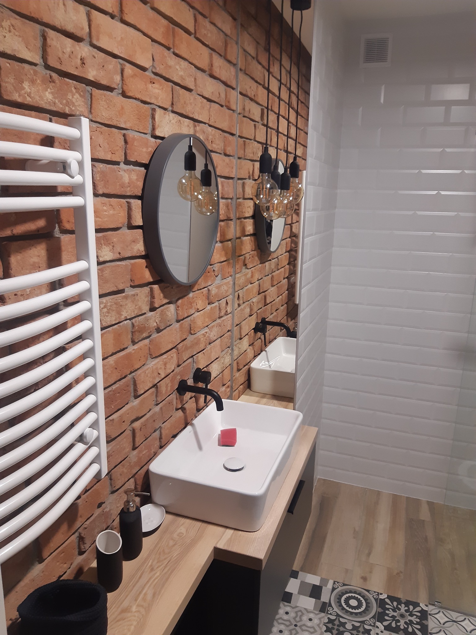 Nowoczesna łazienka z ceglaną ścianą, okrągłym lustrem i wiszącymi żarówkami Edison, biała umywalka na drewnianym blacie i biały grzejnik drabinkowy.