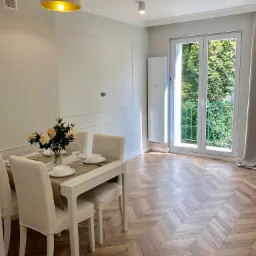 Jasne wnętrze jadalni z parkietem w jodełkę, białym stołem nakrytym do posiłku, z widokiem na balkon z zieloną balustradą i bujną roślinnością za oknem.