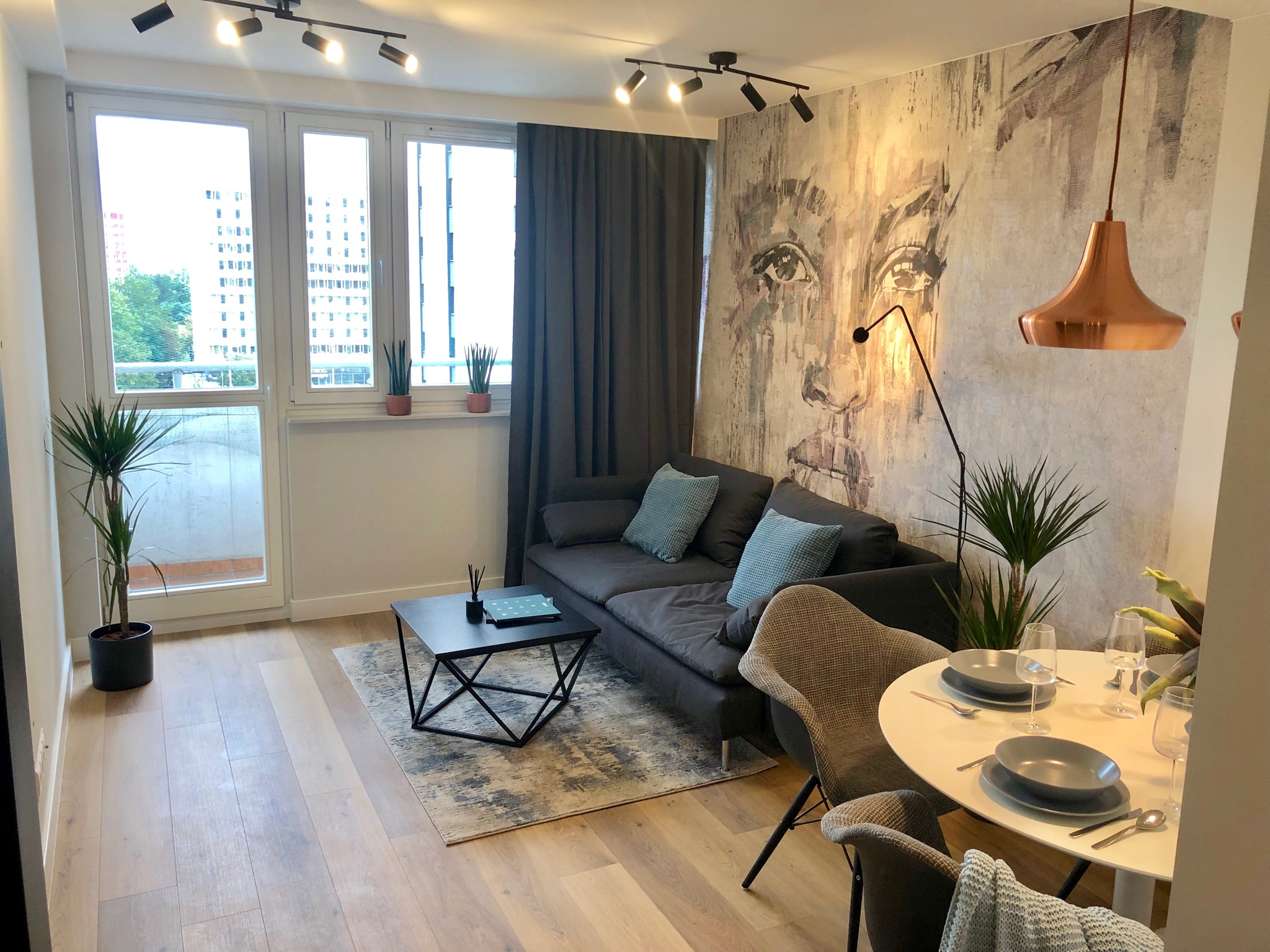 Jasne wnętrze mieszkania z drewnianą podłogą, szarą sofą z turkusowymi poduszkami, okrągłym stołem z nakryciem i ścianą z abstrakcyjnym portretem kobiety.