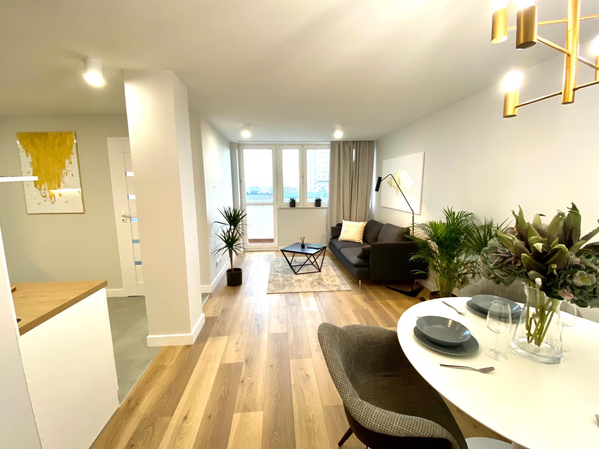 Jasne wnętrze mieszkania po remoncie z drewnianą podłogą, szarą sofą, okrągłym stołem z zastawą i złotą lampą wiszącą.