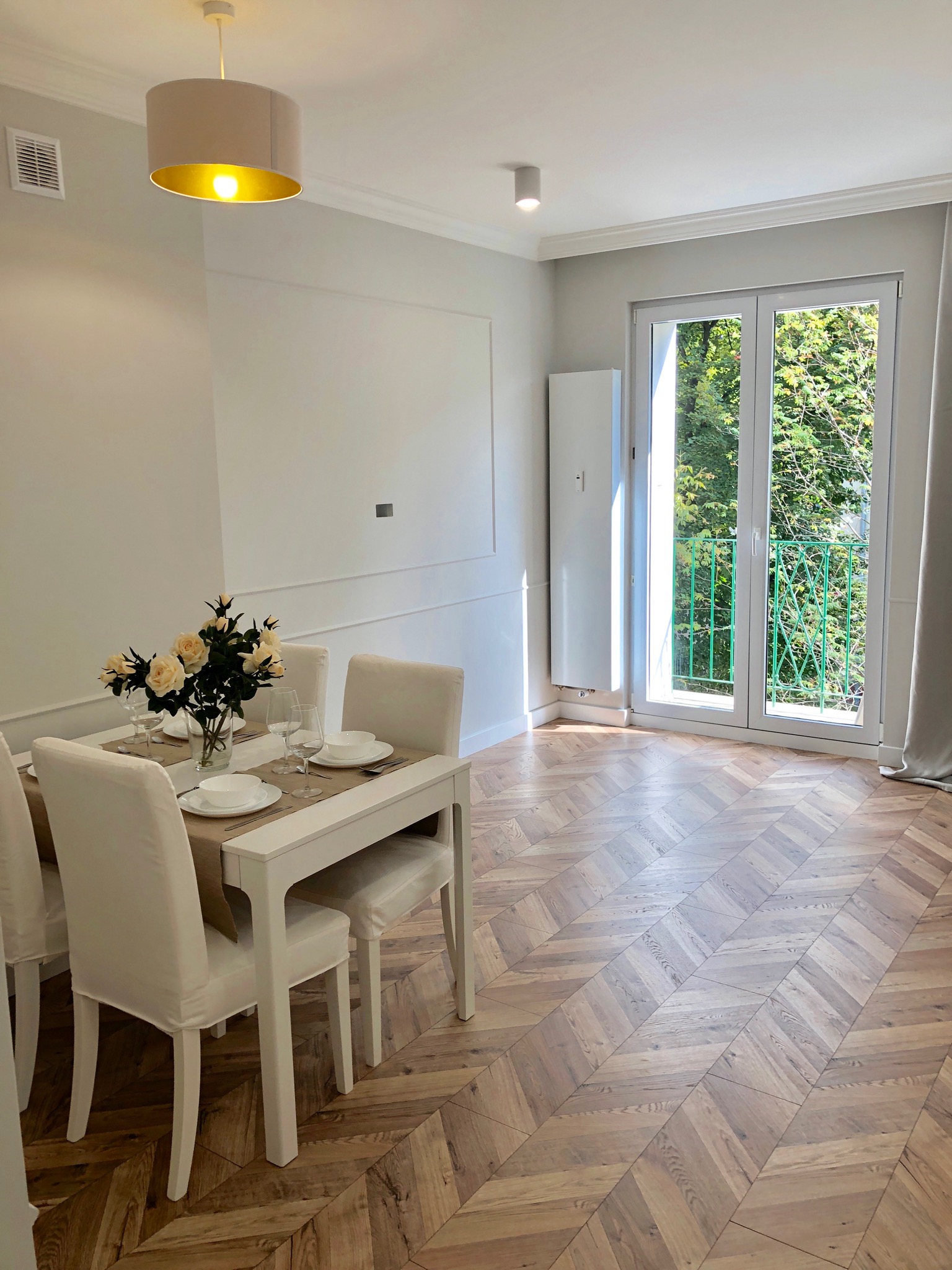 Jasne wnętrze jadalni z parkietem w jodełkę, białym stołem nakrytym do posiłku, z widokiem na balkon z zieloną balustradą i bujną roślinnością za oknem.