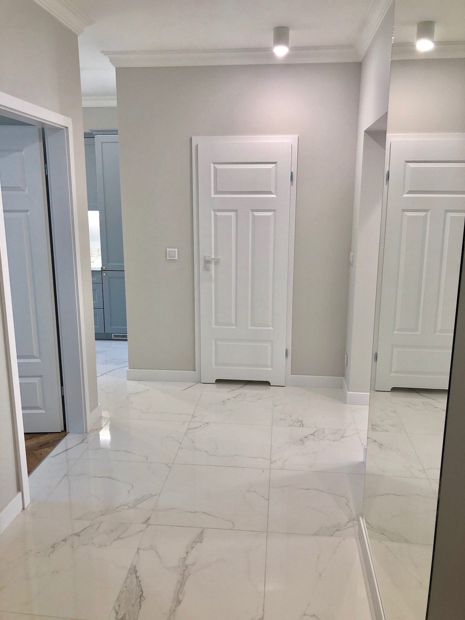 Korytarz w jasnych barwach z białymi drzwiami i marmurową podłogą, widoczne listwy przypodłogowe i oświetlenie sufitowe.