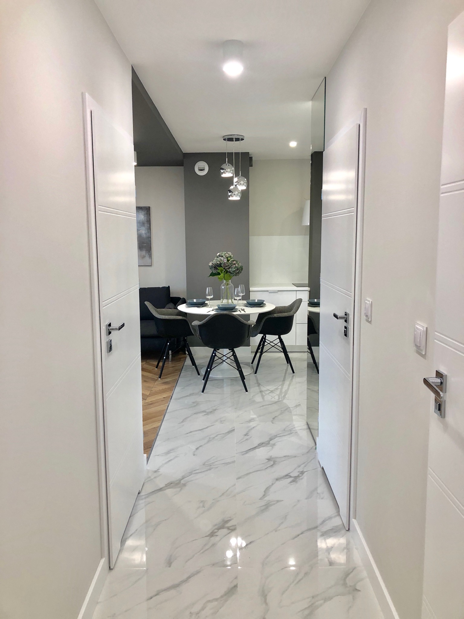 Nowoczesne wnętrze mieszkania z marmurową podłogą, widok z korytarza na jadalnię z okrągłym stołem i szarymi krzesłami oraz salon z szarą sofą w tle.