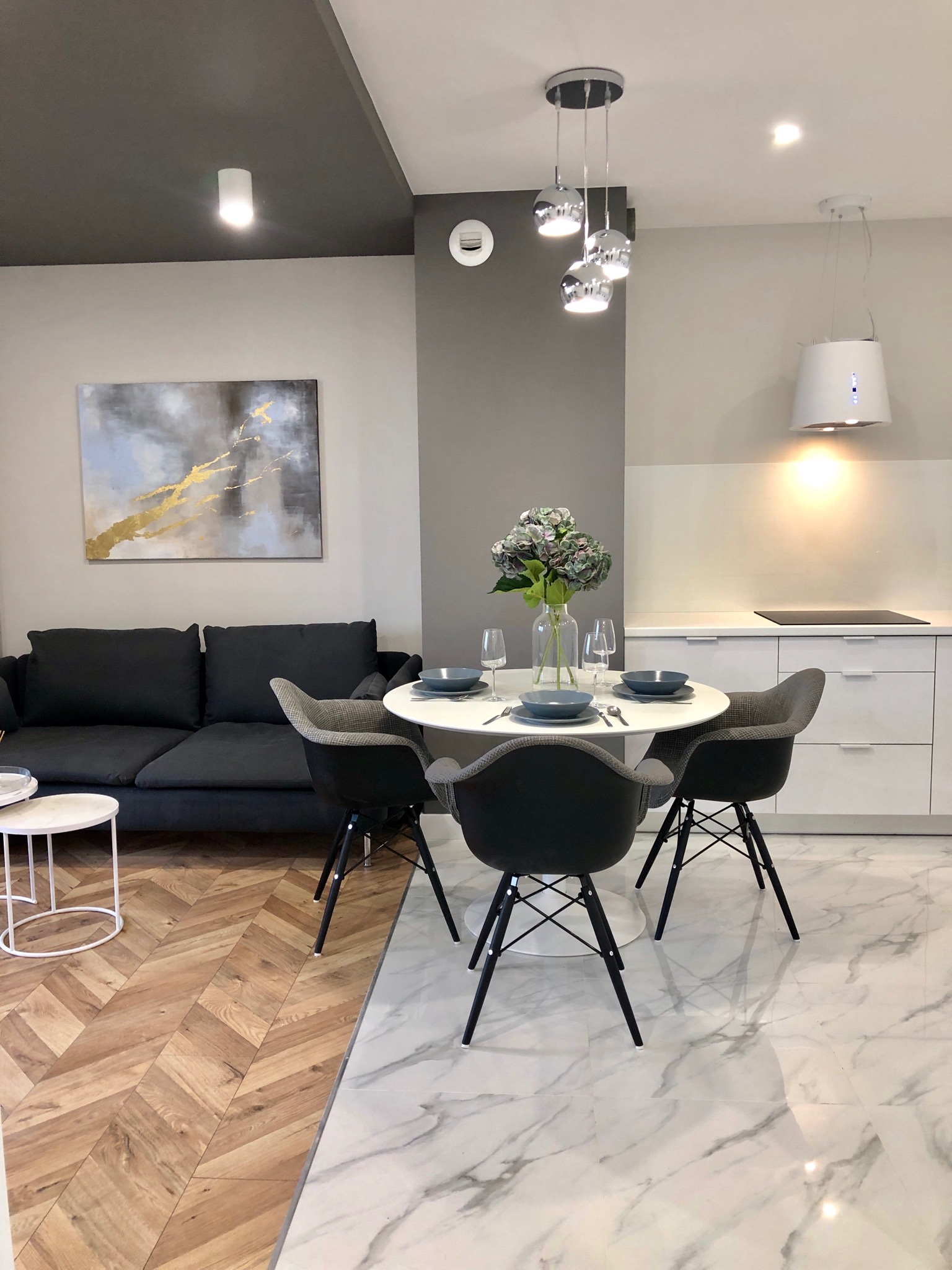Nowoczesne wnętrze: szara sofa, okrągły stół z nakryciem i bukietem hortensji, marmurowa podłoga przechodząca w parkiet w jodełkę, oświetlenie punktowe i lampa wisząca z chromowanymi kulami.