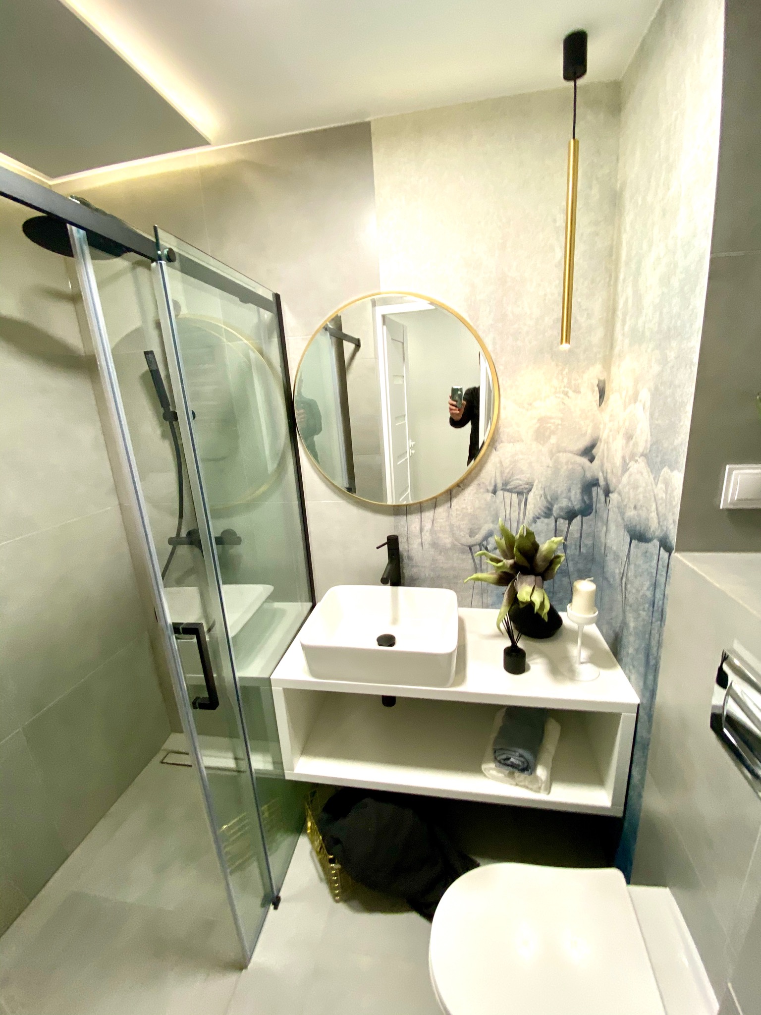 Nowoczesna łazienka z kabiną prysznicową ze szklanymi drzwiami z czarnymi ramami, okrągłym lustrem w złotej ramie, białą umywalką nablatową na szafce i tapetą w delikatne wzory roślinne.