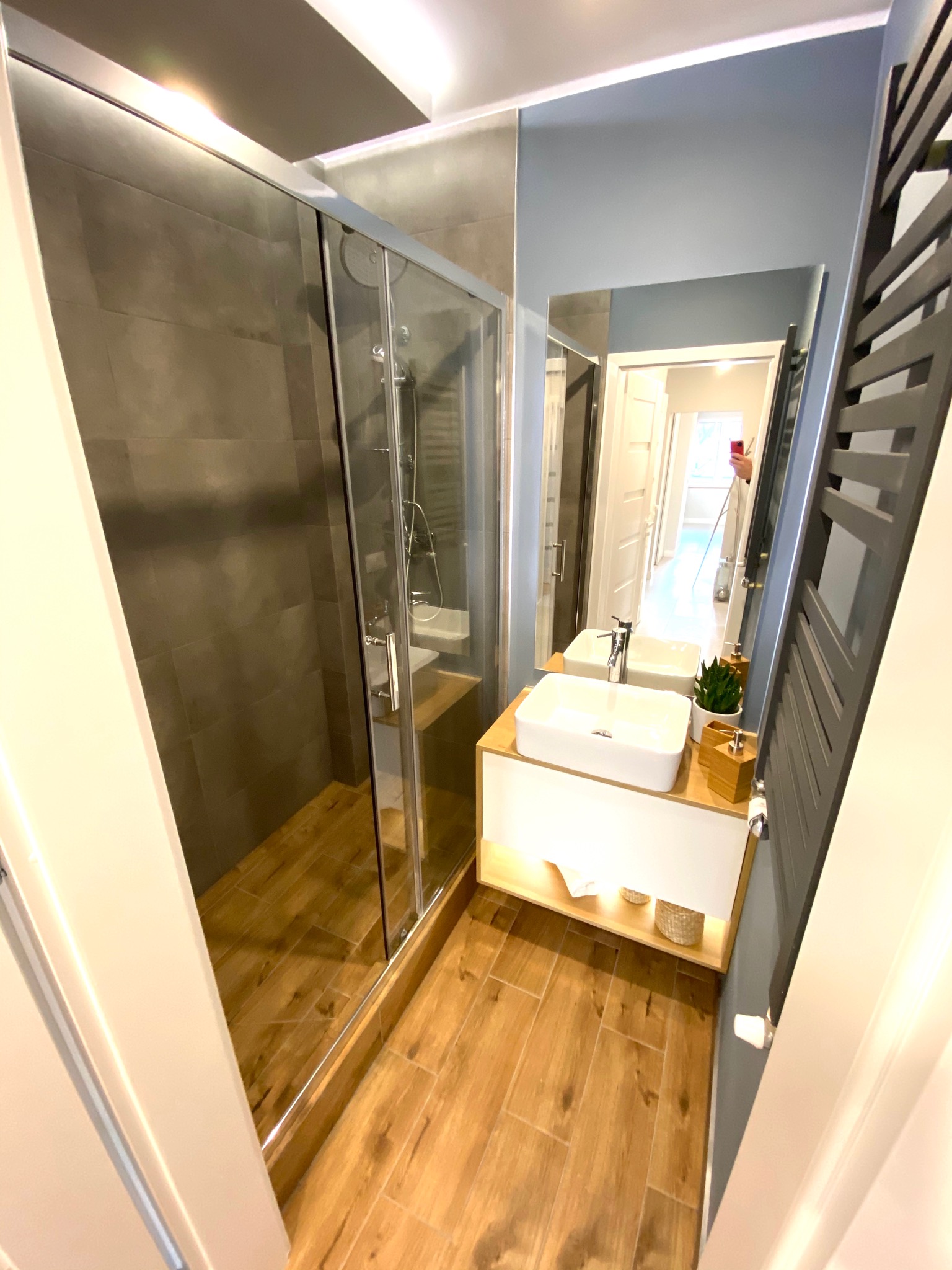 Nowoczesna łazienka z kabiną prysznicową z ciemnymi płytkami, drewnianymi panelami podłogowymi i białą umywalką na podświetlanej szafce, widziana z perspektywy wejścia.