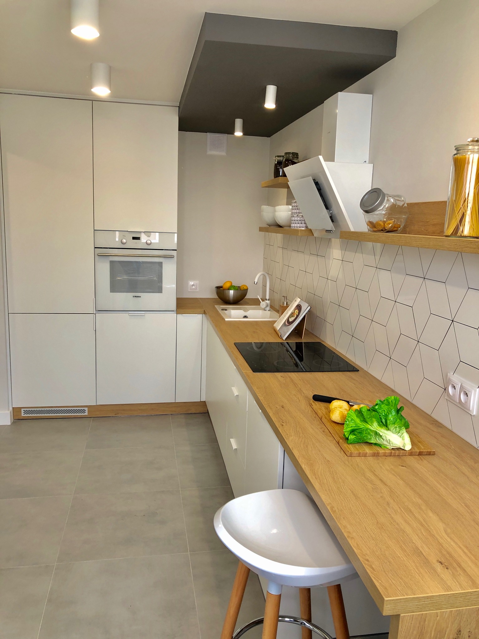 Jasna kuchnia z białymi szafkami, drewnianym blatem i geometrycznymi płytkami na ścianie, na blacie leżą warzywa na desce do krojenia, nad blatem półki z dekoracjami i produktami spożywczymi