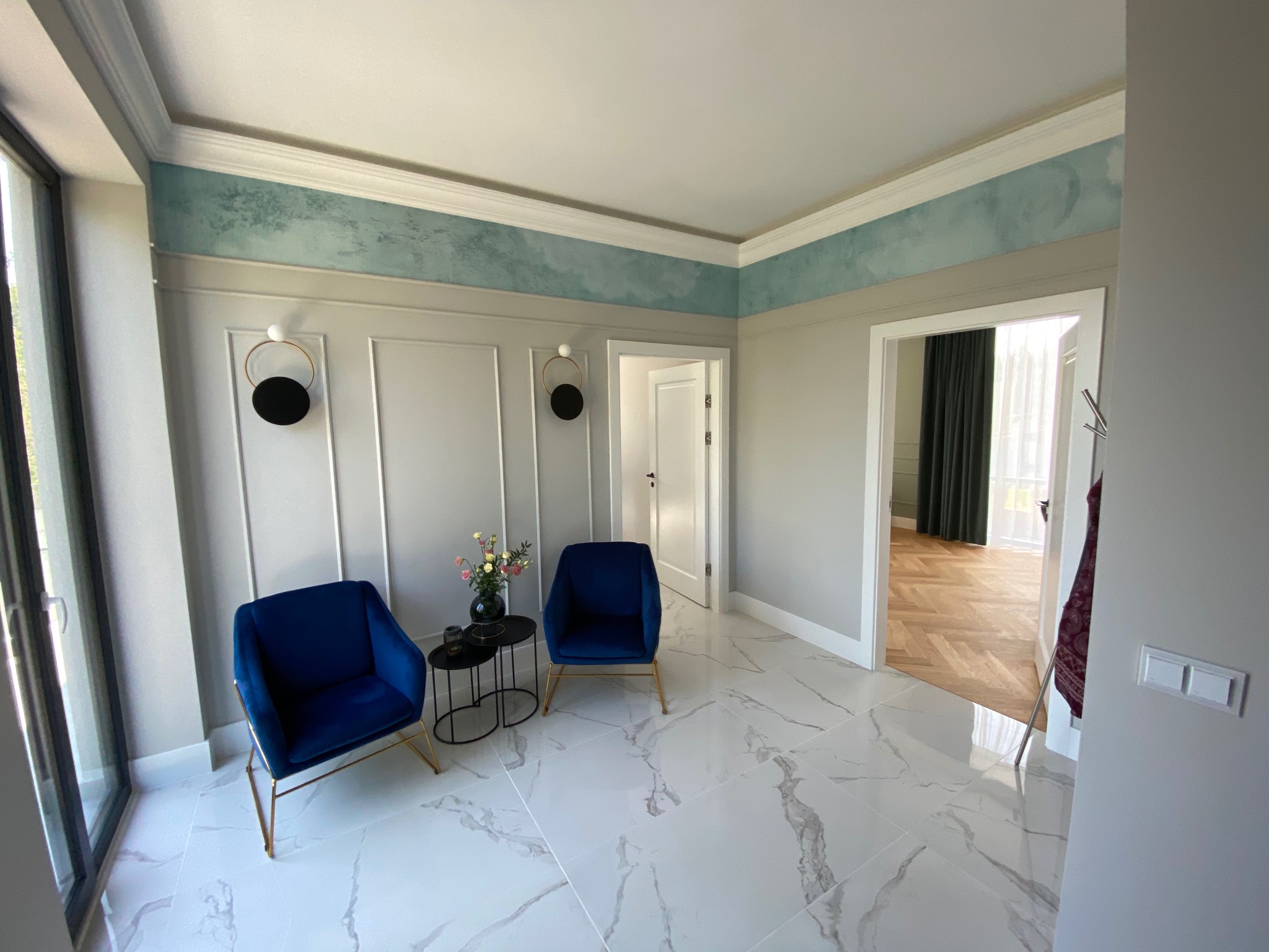 Elegancki hol z dwiema granatowymi, welurowymi fotelami na złotych nóżkach, marmurową podłogą z szarymi żyłami, dekoracyjną ścianą z panelami i abstrakcyjną malaturą w odcieniach błękitu pod sufitem.
