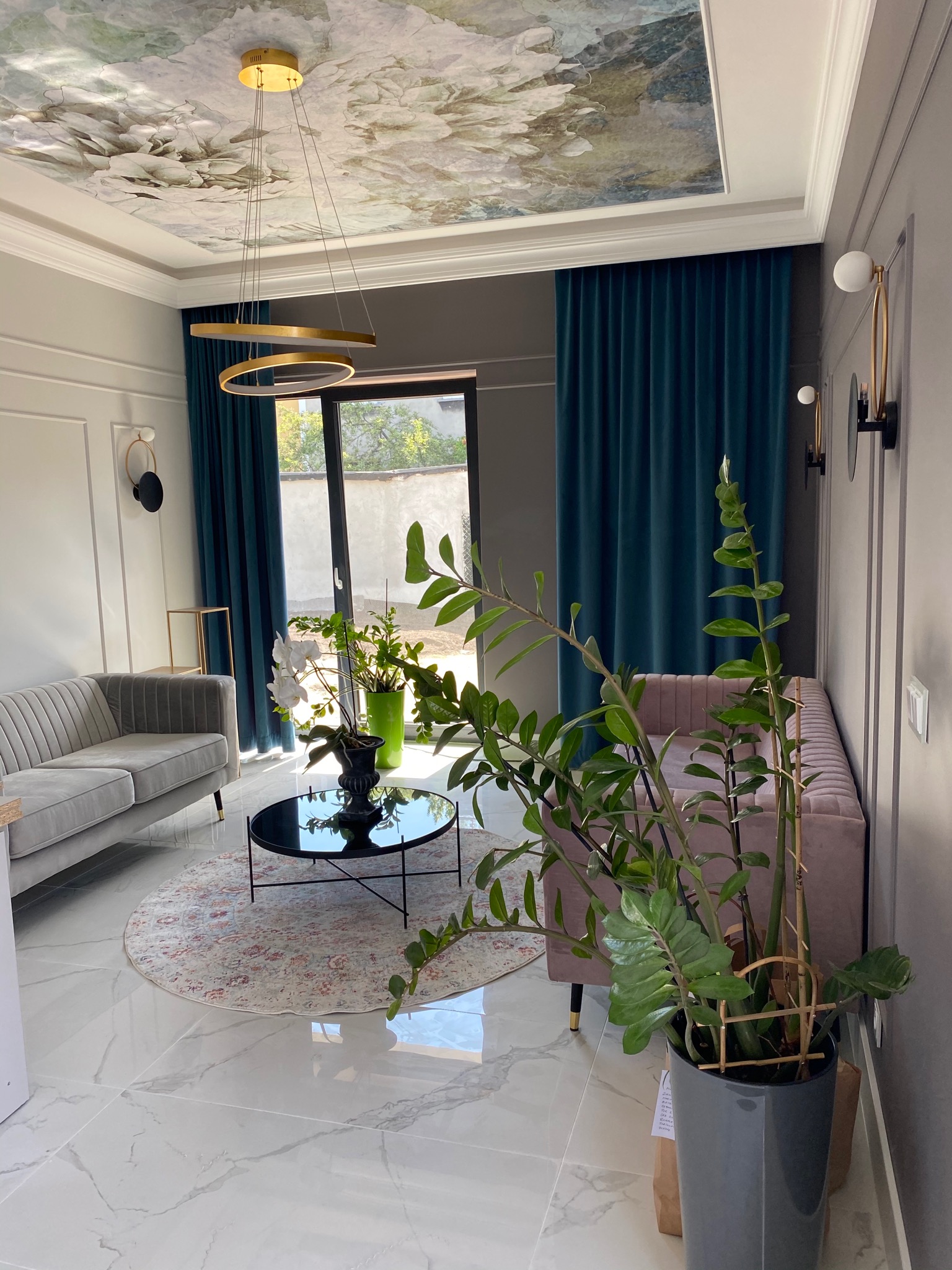 Elegancki salon z szarymi i różowymi sofami, okrągłym dywanem i czarnym stolikiem kawowym, ozdobiony kwiatami, roślinami doniczkowymi, turkusowymi zasłonami i dekoracyjnym malowaniem sufitu.