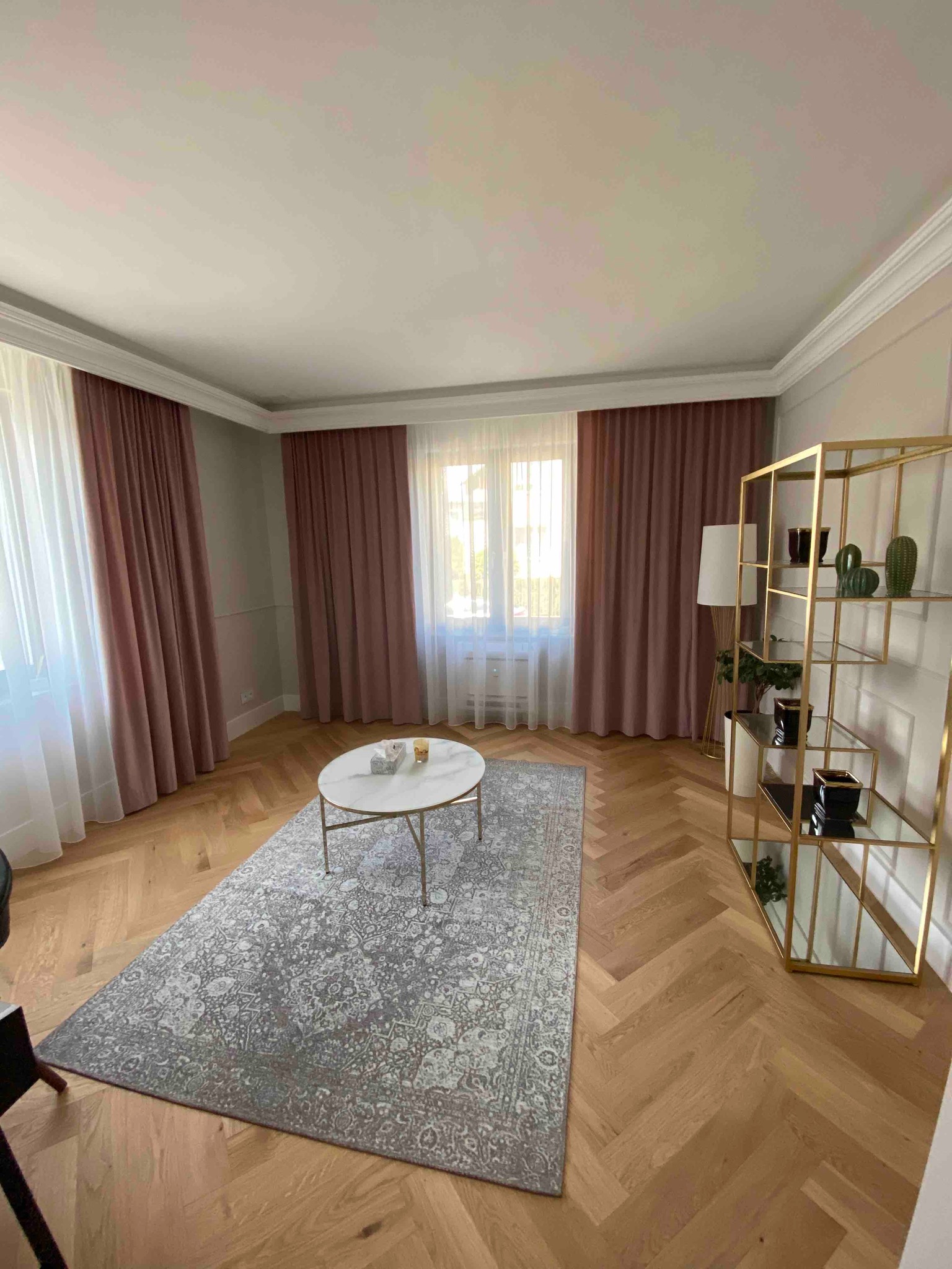 Jasny salon z parkietem w jodełkę, marmurowym stolikiem kawowym, wzorzystym dywanem i różowymi zasłonami.