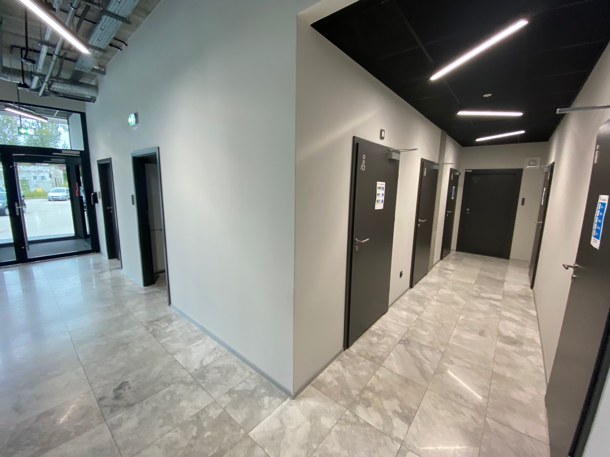 Korytarz biurowy z szarymi ścianami, ciemnymi drzwiami i marmurową podłogą, widoczne instalacje pod sufitem po lewej stronie, nowoczesne oświetlenie sufitowe po prawej.