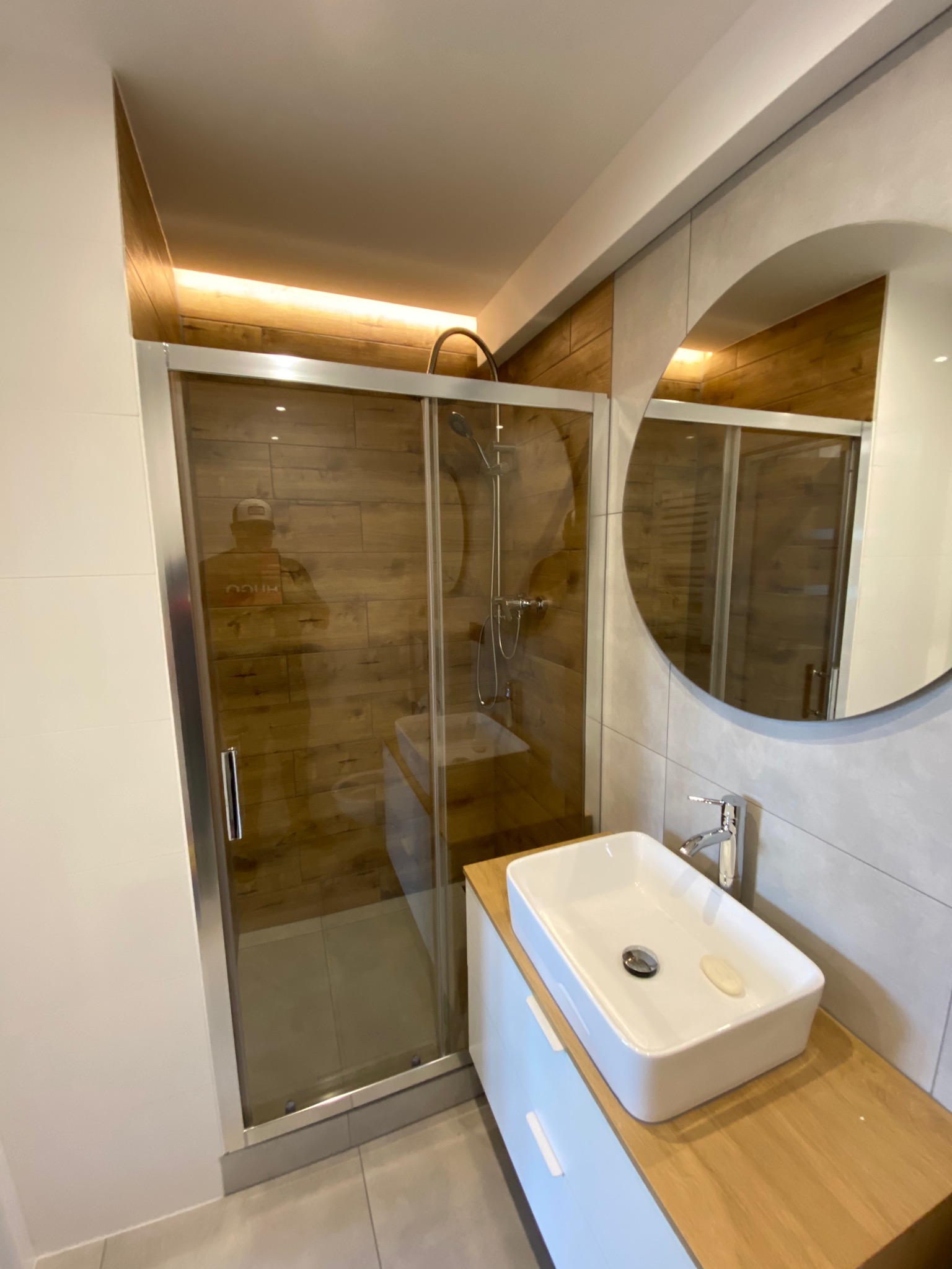 Nowoczesna łazienka z kabiną prysznicową ze szklanymi drzwiami przesuwnymi, umywalką nablatową na drewnianym blacie, okrągłym lustrem i oświetleniem LED w suficie.