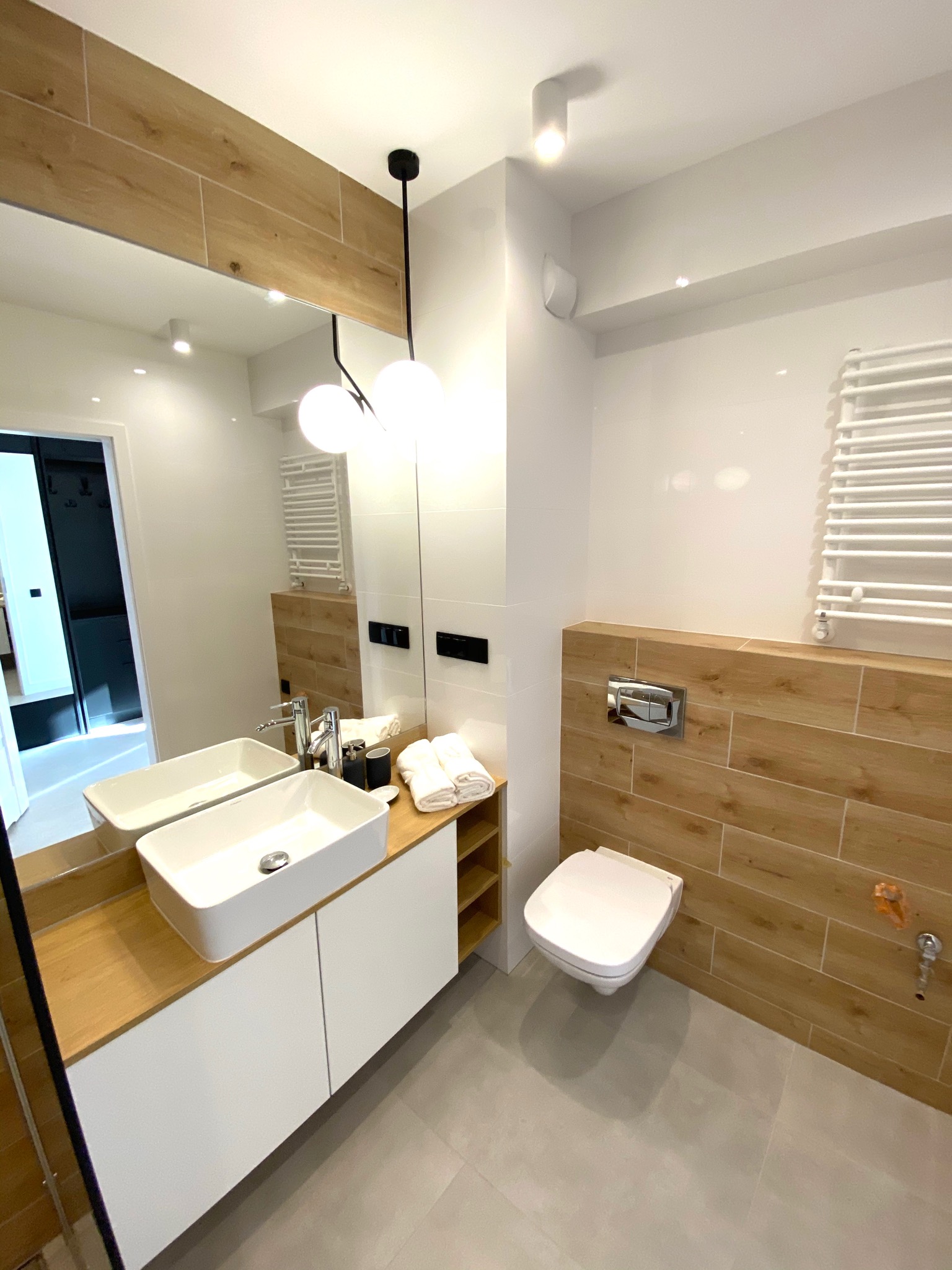 Nowoczesna łazienka z białymi płytkami, drewnopodobnymi akcentami, kwadratową umywalką na białej szafce i wiszącą toaletą; widoczne oświetlenie sufitowe i grzejnik drabinkowy.