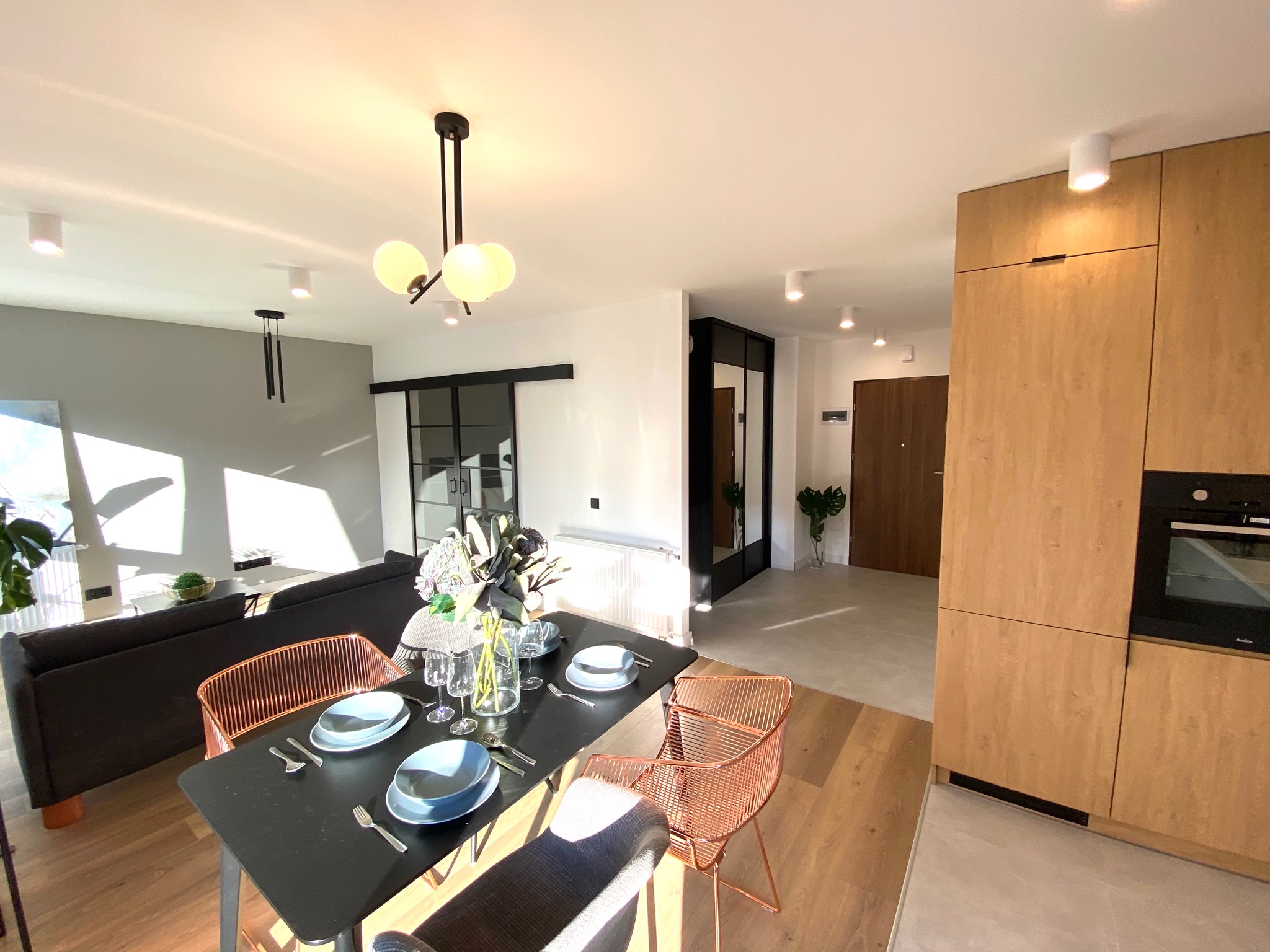 Jasne, przestronne wnętrze mieszkania z otwartą kuchnią, jadalnią i salonem. Widoczny czarny stół z nakryciem, miedziane krzesła i nowoczesna lampa wisząca. W tle sofa i przesuwne drzwi.