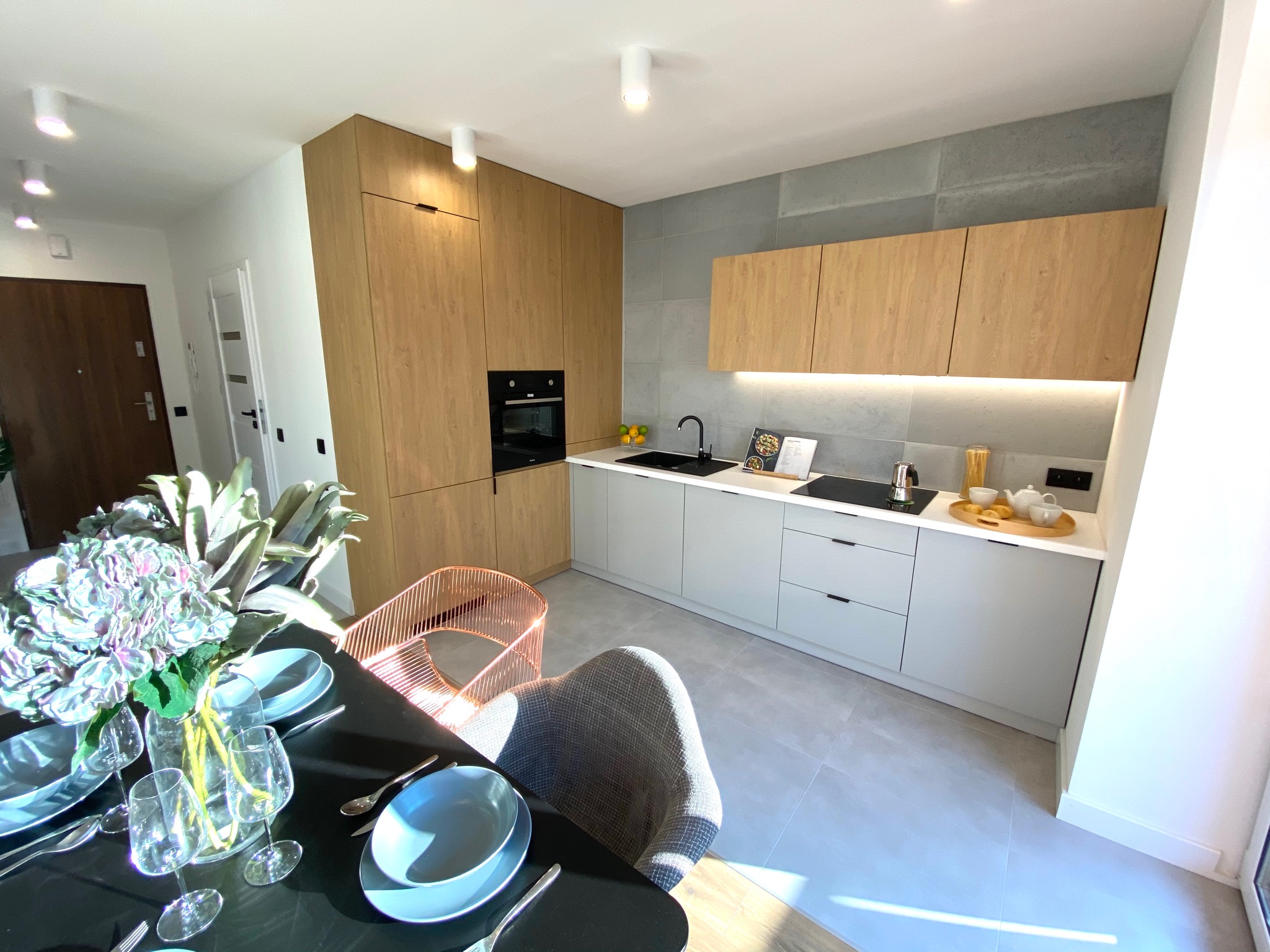 Nowoczesna kuchnia z drewnianymi szafkami, szarymi płytkami na ścianie i czarnym zlewem, widok z jadalni z nakrytym stołem i kwiatami w wazonie.