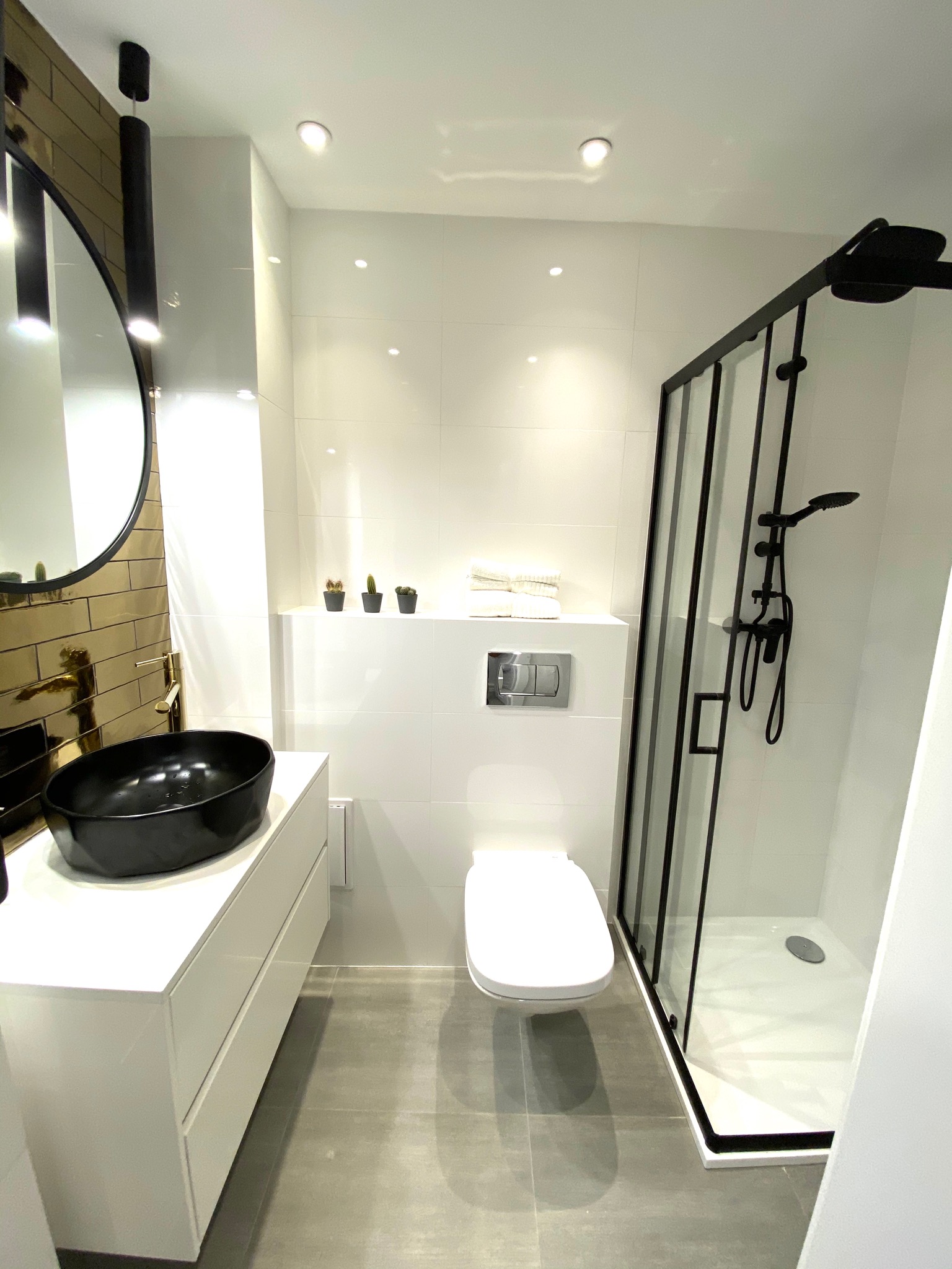 Nowoczesna łazienka z czarną umywalką nablatową, złotą armaturą i kabiną prysznicową z czarnymi ramami, doświetlona punktowymi lampami sufitowymi.