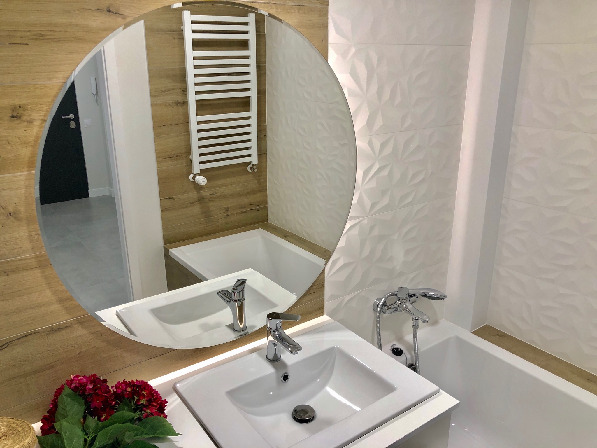 Nowoczesna łazienka z okrągłym lustrem, białymi płytkami 3D i drewnopodobną okładziną, widoczny fragment umywalki, w odbiciu grzejnik drabinkowy.