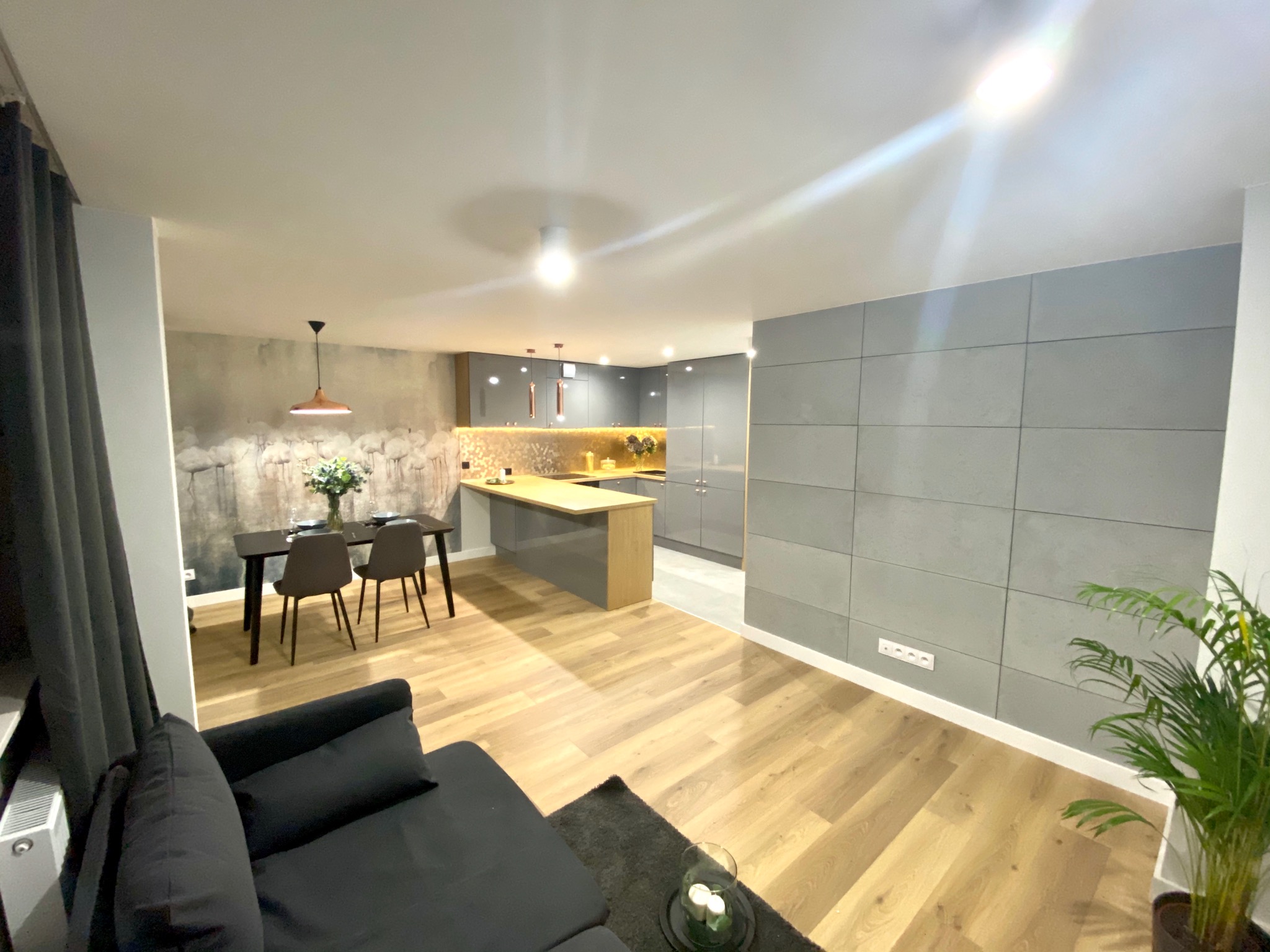 Nowoczesne wnętrze mieszkania z otwartą kuchnią, drewnianą podłogą, szarymi meblami kuchennymi i ścianą z płyt betonowych. W tle widoczny stół z krzesłami i dekoracyjna tapeta.