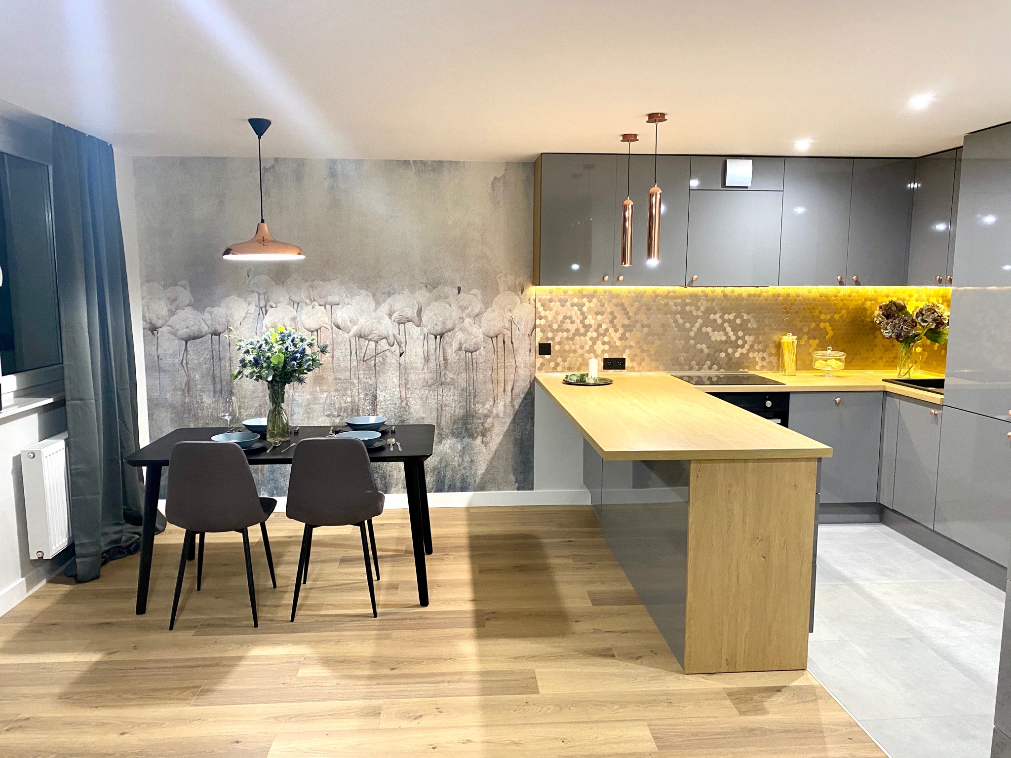 Nowoczesna kuchnia z szarymi, lakierowanymi szafkami, drewnianym blatem i heksagonalną mozaiką nad blatem, połączona z jadalnią z czarnym stołem i krzesłami na parkiecie, ozdobiona tapetą z motywem...