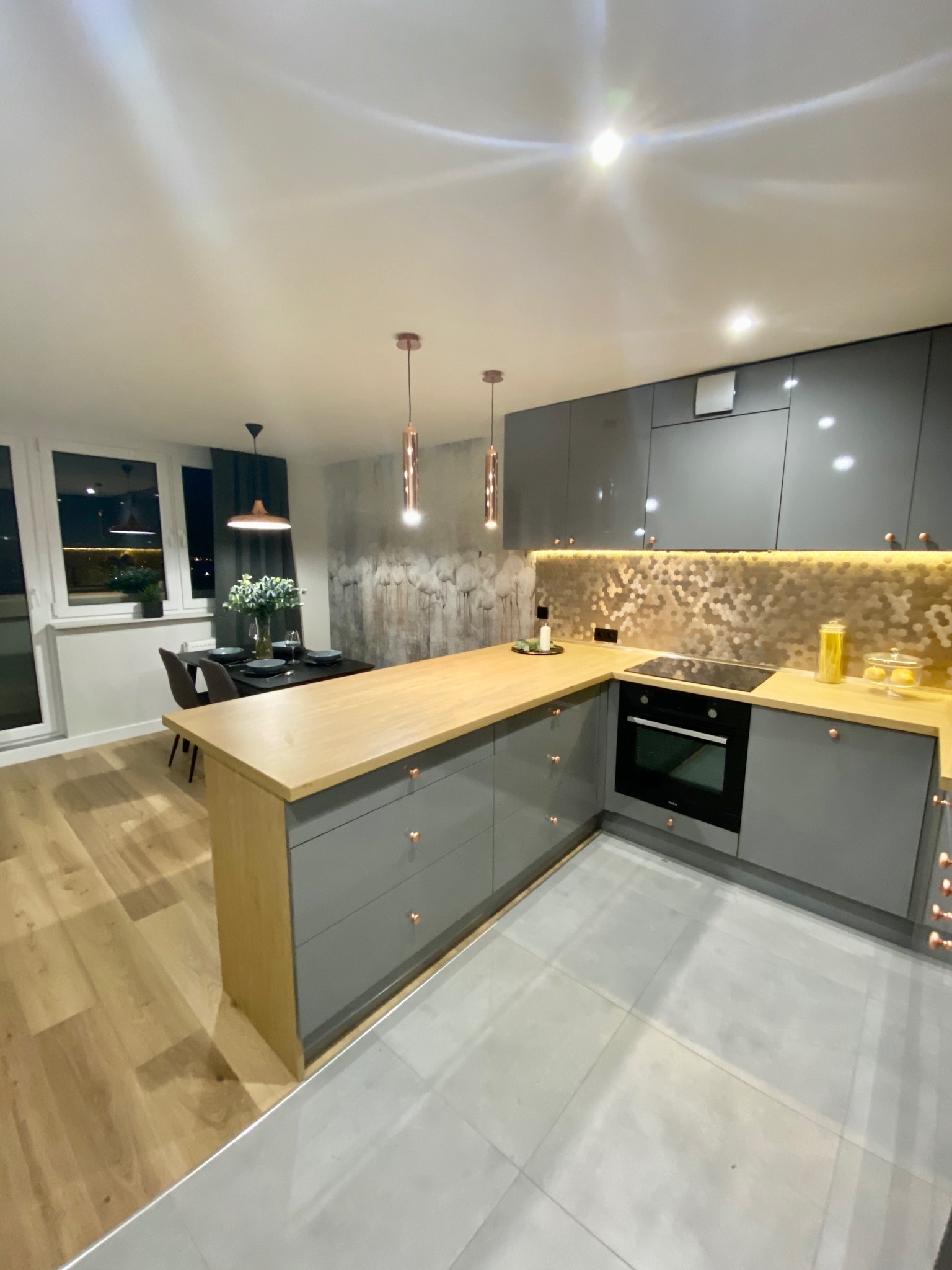 Nowoczesna kuchnia w odcieniach szarości z drewnianym blatem i heksagonalną mozaiką nad blatem, oświetlona wiszącymi lampami w kolorze miedzi i podświetleniem LED.