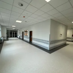 Jasny, przestronny korytarz w budynku użyteczności publicznej z białymi ścianami, szarymi pasami dekoracyjnymi, panelami sufitowymi i jasną podłogą, widoczne okna po lewej stronie.