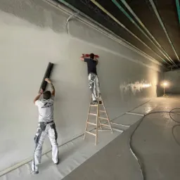 Dwóch pracowników w trakcie wygładzania ściany w dużym pomieszczeniu; jeden stoi na drabinie, drugi na podłodze, obaj w roboczych ubraniach; podłoga zabezpieczona folią, widoczne oświetlenie...