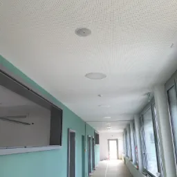 Długi korytarz biurowy z perforowanym sufitem podwieszanym i wbudowanymi okrągłymi lampami, ściany pomalowane na pastelowy turkus, podłoga zabezpieczona folią malarską.