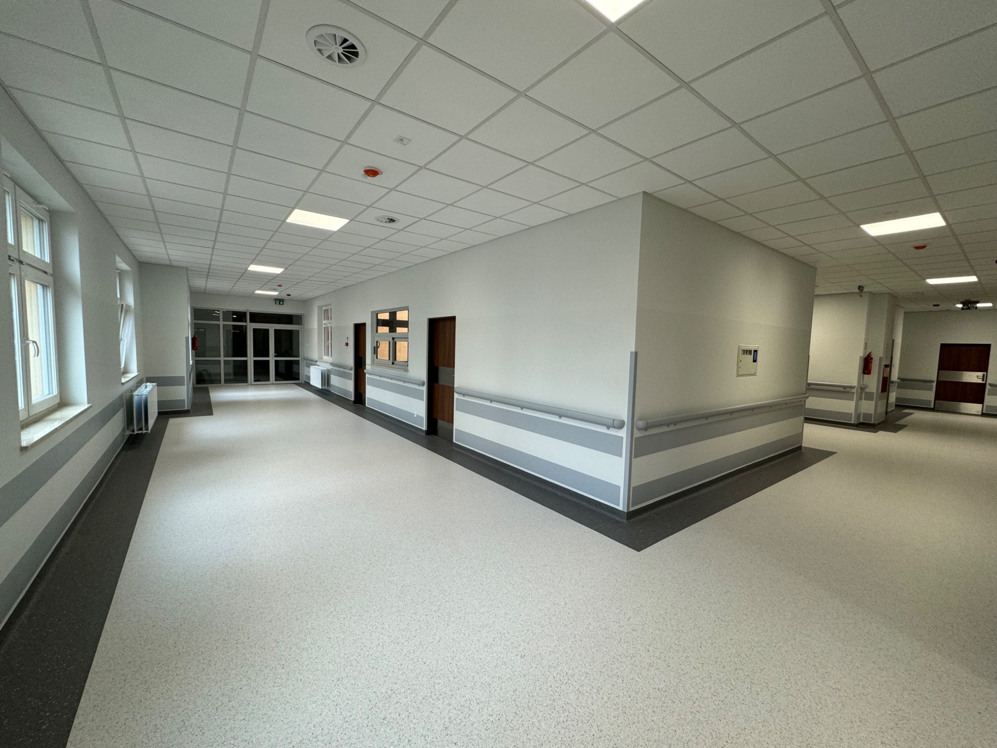Jasny, przestronny korytarz w budynku użyteczności publicznej z białymi ścianami, szarymi pasami dekoracyjnymi, panelami sufitowymi i jasną podłogą, widoczne okna po lewej stronie.