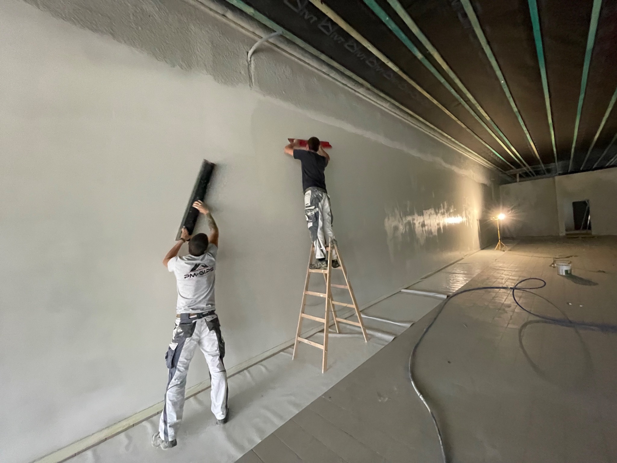 Dwóch pracowników w trakcie wygładzania ściany w dużym pomieszczeniu; jeden stoi na drabinie, drugi na podłodze, obaj w roboczych ubraniach; podłoga zabezpieczona folią, widoczne oświetlenie...