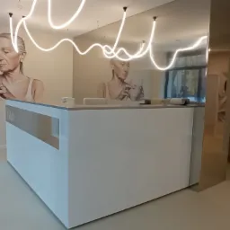 Biała lada recepcyjna z logo 'Gaia' w nowoczesnym wnętrzu, dekorowana muralem z motywem kobiecej twarzy i oświetleniem neonowym.