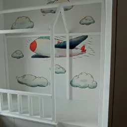 Biała konstrukcja przypominająca domek, prawdopodobnie rama łóżka dziecięcego, z naklejkami przedstawiającymi samolot i chmury, umieszczona w rogu pokoju obok okna i ciemnej szafy.