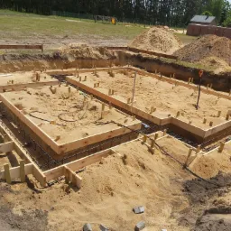 Przygotowanie fundamentów pod dom murowany: drewniane szalunki zbrojone stalą, wypełnione piaskiem, widoczne wykopane fundamenty i hałdy ziemi w tle.