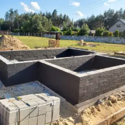 Fundamenty z bloczków betonowych zabezpieczone izolacją przeciwwilgociową, widoczny stos bloczków i usypany piasek, w tle dom jednorodzinny i las.