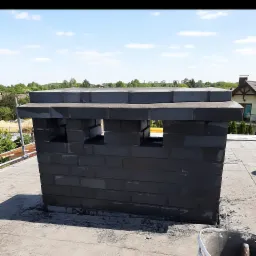 Murowany komin z czarnej cegły na płaskim dachu, z widocznymi otworami wentylacyjnymi, na tle błękitnego nieba i zieleni drzew.