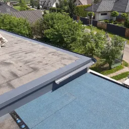 Widok z góry na fragment dachu płaskiego z widocznym połączeniem starej i nowej hydroizolacji, z blachy okapowej i z fragmentu miasta w tle.