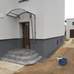 Wykończona elewacja domu z szarym cokołem, wejściem z zadaszeniem i schodami wyłożonymi płytkami, widoczne wiadro i kuweta malarska.