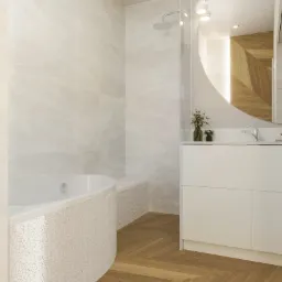 Jasna łazienka z wanną obudowaną mozaiką, drewnianą podłogą w jodełkę, marmurowymi płytkami ściennymi i okrągłym lustrem z podświetleniem LED.