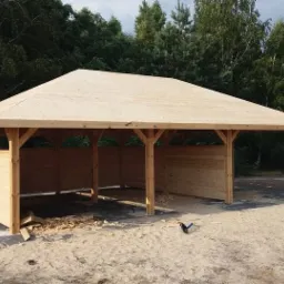 Surowa drewniana altana z płaskim dachem, częściowo obudowana deskami, widoczna na tle zieleni, z drobnymi odpadami po obróbce drewna wokół.