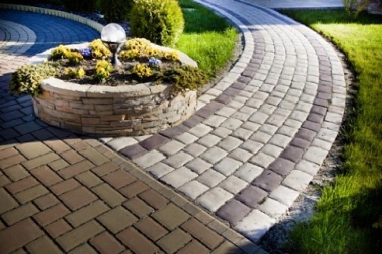 Ogród z wybrukowaną ścieżką z kostki brukowej w dwóch odcieniach szarości, obok okrągła rabata z kwiatami i lampą, trawnik w tle.