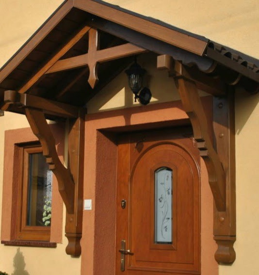 Drewniany daszek nad wejściem do domu z ozdobnymi wspornikami i stylizowaną latarnią, brązowa stolarka okienna i drzwiowa.